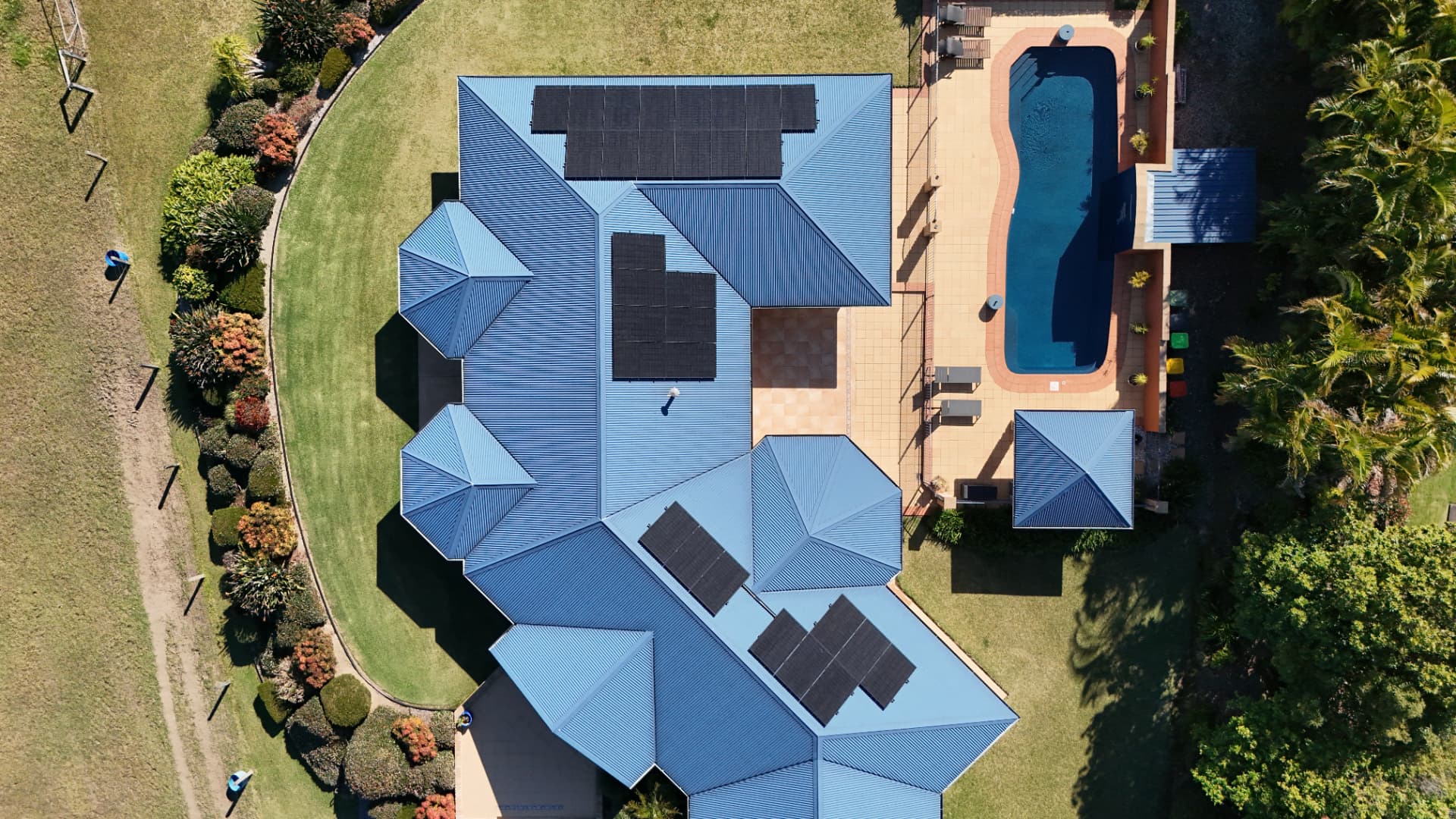 Aerial view of a Coffs Harbour home with black solar panels across its blue tiled roof beside a pool