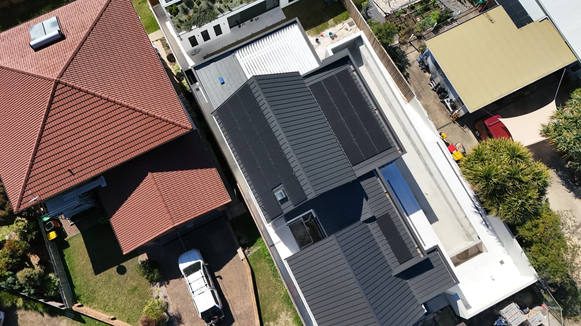 Overhead drone view of a modern dark grey metal roof fitted with a full black solar array, beside a neighbouring tiled-roof home